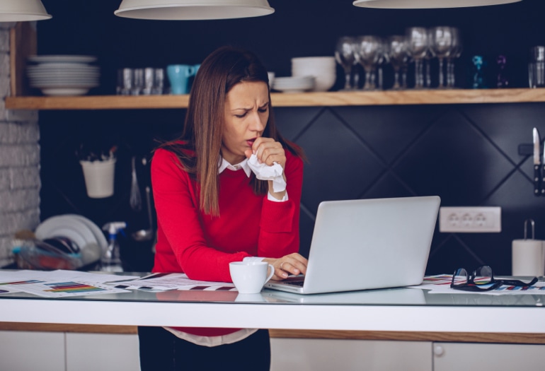 Co powoduje kaszel? Kobieta stoi w kuchni i pracuje na laptopie, kaszląc chusteczkę.